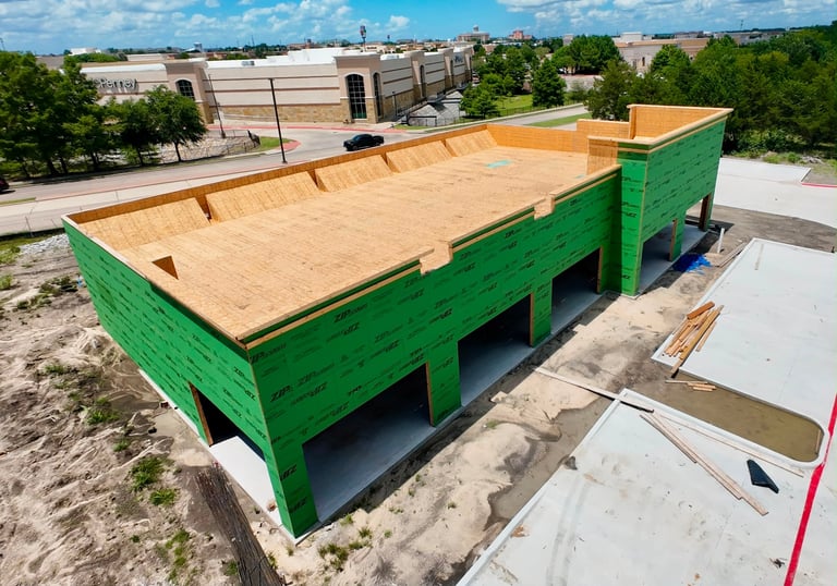 Large green wooden agricultural trailer with plywood bed on construction site with warehouses in background
