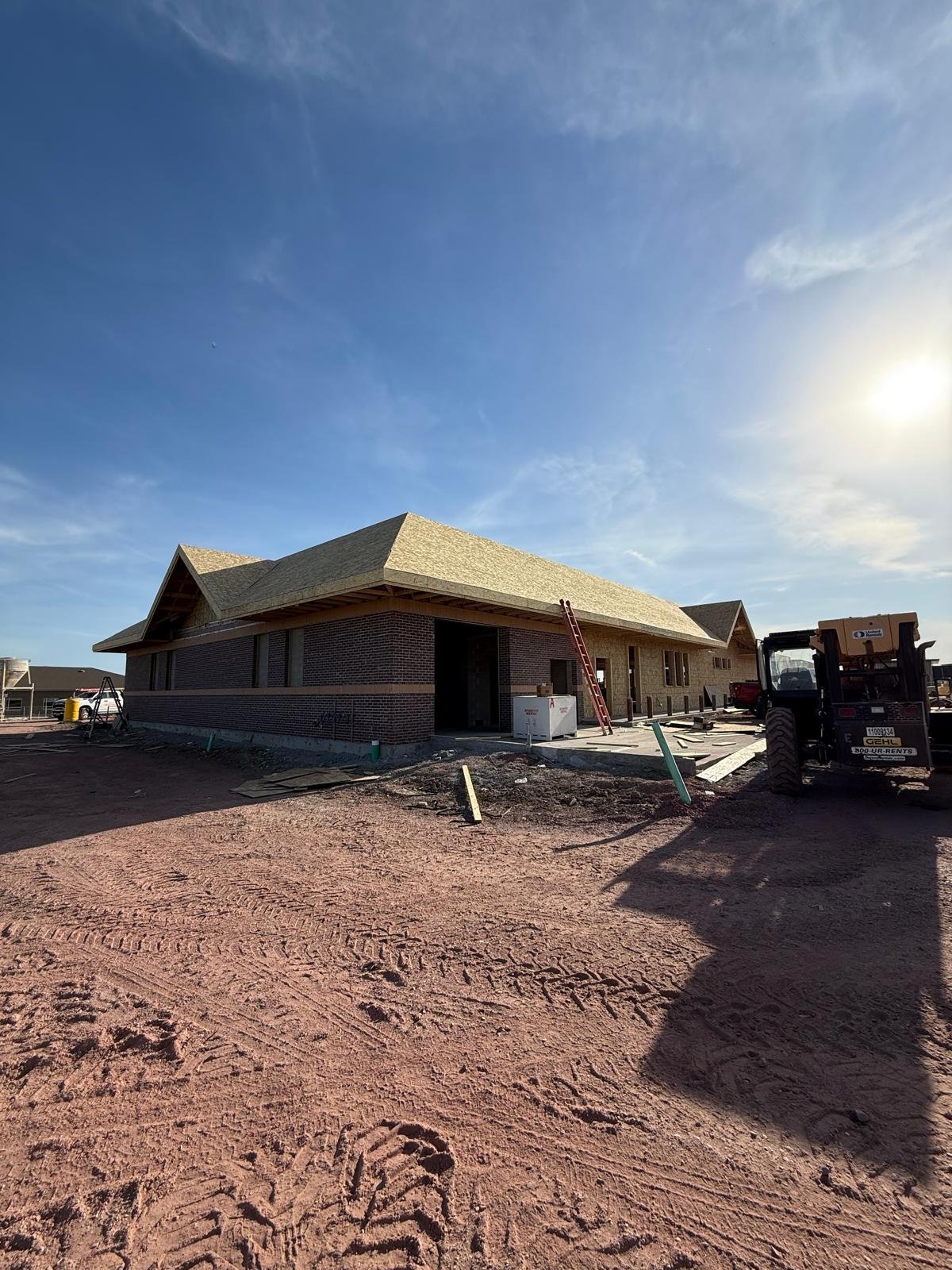 New residential house under construction with shingled roof, brick exterior, and construction equipment on sandy ground under blue sky