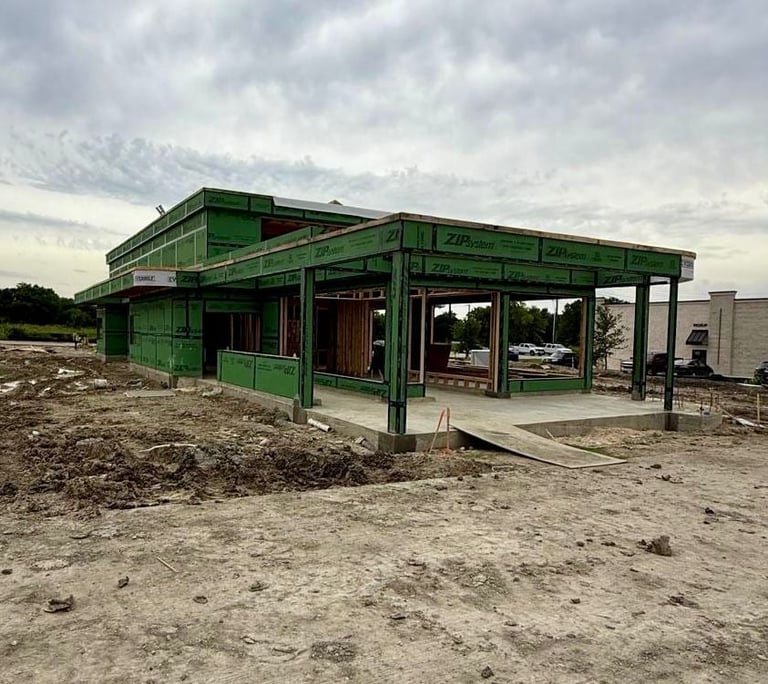 Green metal frame commercial building under construction on bare dirt lot with mountains in background