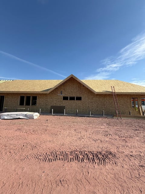 Brick building under construction with new shingle roof, wooden ladder, and red clay ground against blue sky