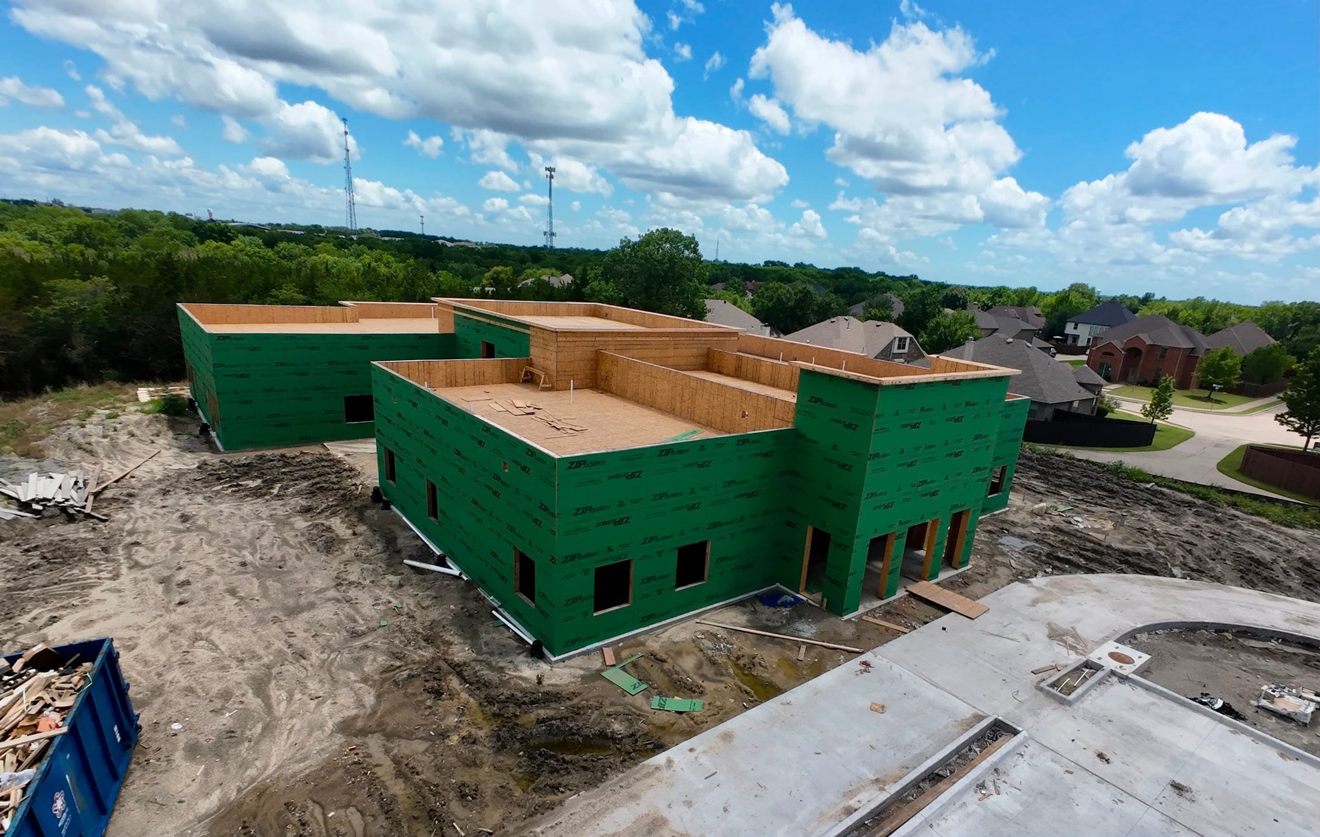 Aerial view of a green commercial building under construction with wooden roof framing and exposed interior walls on a cleared lot.