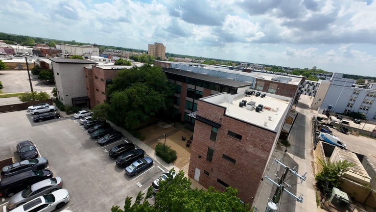 Aerial view of an industrial complex with brick and metal buildings, parked vehicles, and green vegetation under a partly cloudy sky