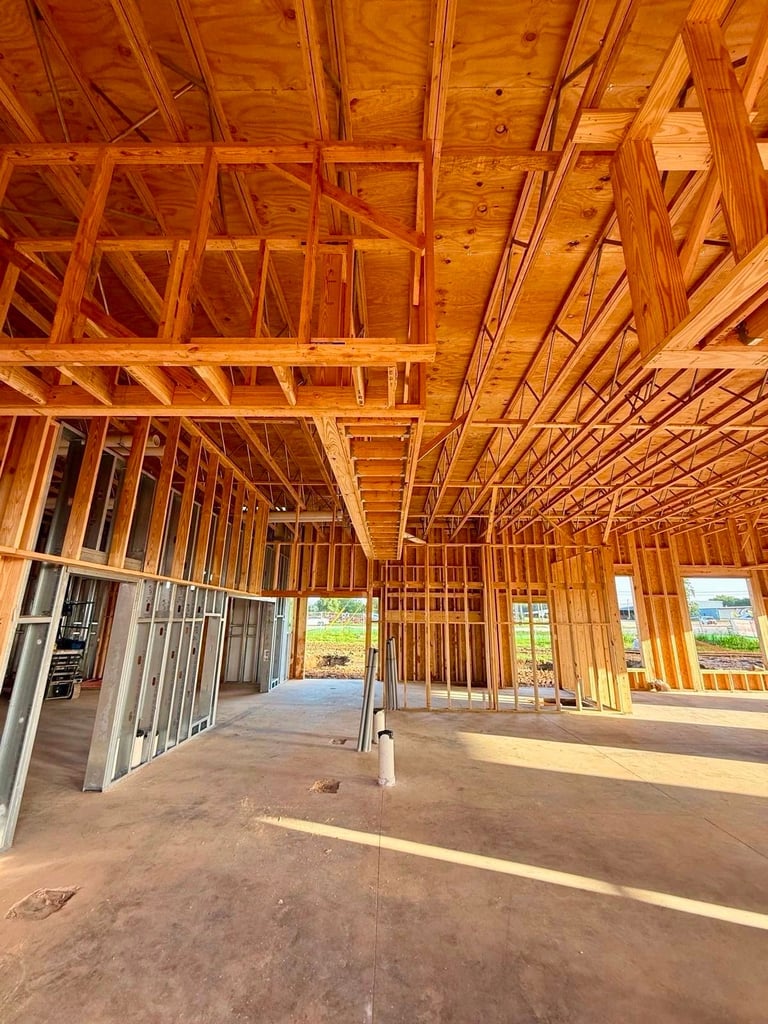 Interior of a wooden-framed building under construction with exposed beams, concrete floor, and windows showing rural landscape outside