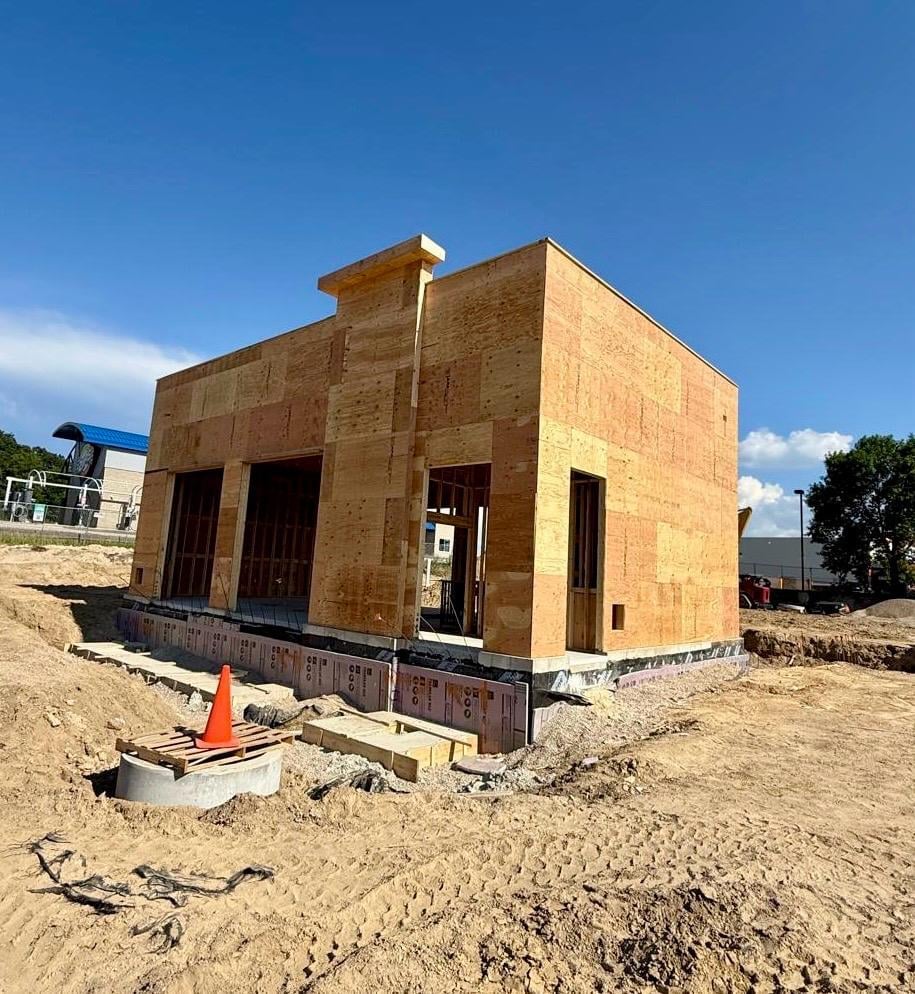 A brick house under construction on bare ground with an orange traffic cone in the foreground and clear blue sky above.