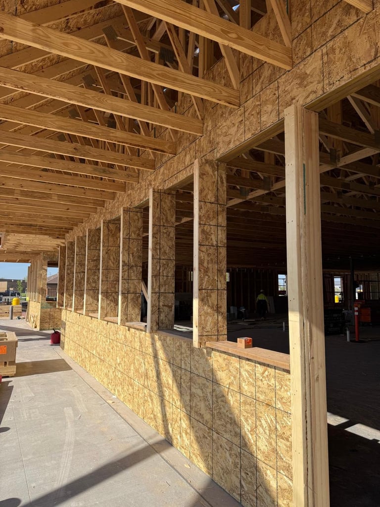 Interior of a building under construction showing exposed wooden beam framing, stone walls, and concrete flooring with large window openings