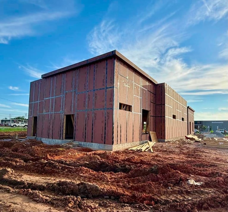 Large industrial-style building under construction on bare dirt lot with clear blue sky, showing wooden frame and metal siding