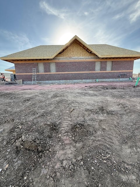 Brick house under construction with new roof framing on unpaved dirt lot under blue sky