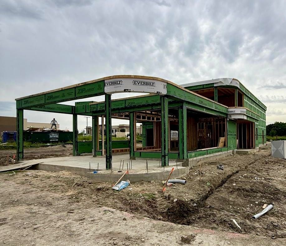 A green metal-frame building under construction on bare dirt ground with overcast sky above