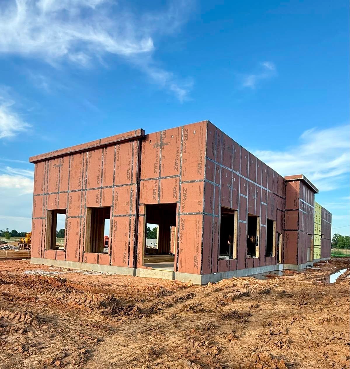 Construction site showing a brick building frame with large window and door openings on bare earth under a blue sky