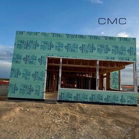Construction house frame with green weather barrier wrap and exposed wooden framing against a blue sky