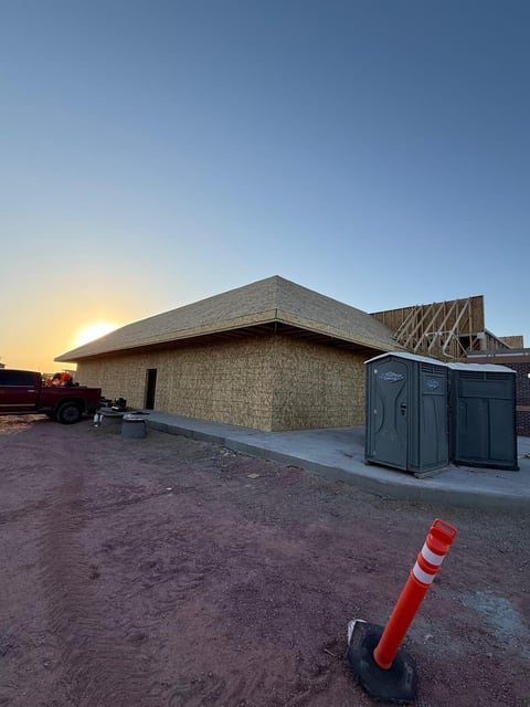 Modern building under construction with straw-colored walls, peaked roof, portable toilets, red and white safety bollard, sunrise setting