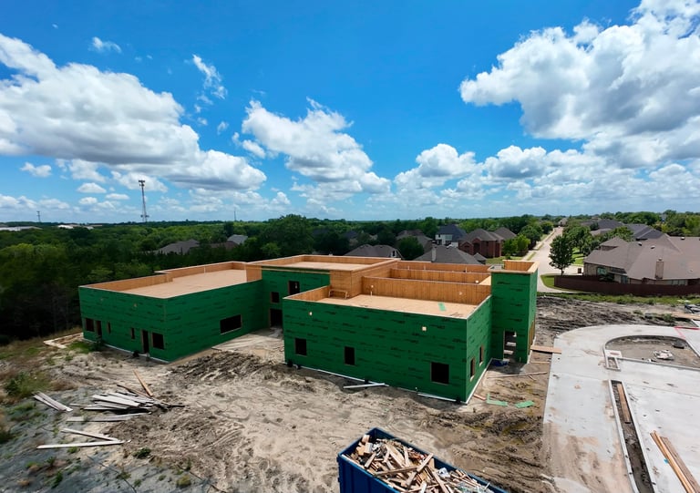 Aerial view of a green commercial building under construction with exposed wooden framing, surrounded by residential homes and trees under blue sky