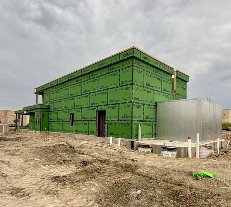 A green residential building under construction with metal framework and sheathing, next to a metal water tank on bare ground