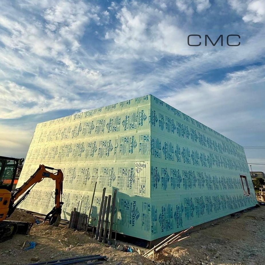 Construction site with large foam insulation-wrapped building structure and yellow excavator nearby under blue cloudy sky
