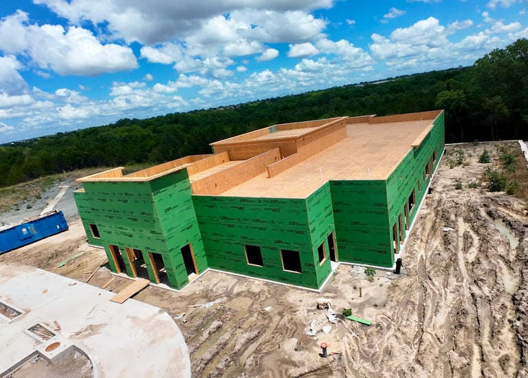 Large commercial building under construction with green exterior walls and wooden roof frame on a cleared site surrounded by forest