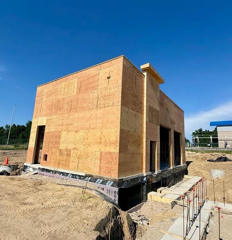 Brick house under construction on sandy lot with foundation, open windows and doorways, and protective fencing.