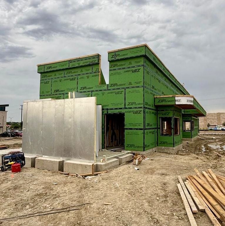 Construction site showing a green-framed house under construction with white metal tank and wooden materials on bare ground
