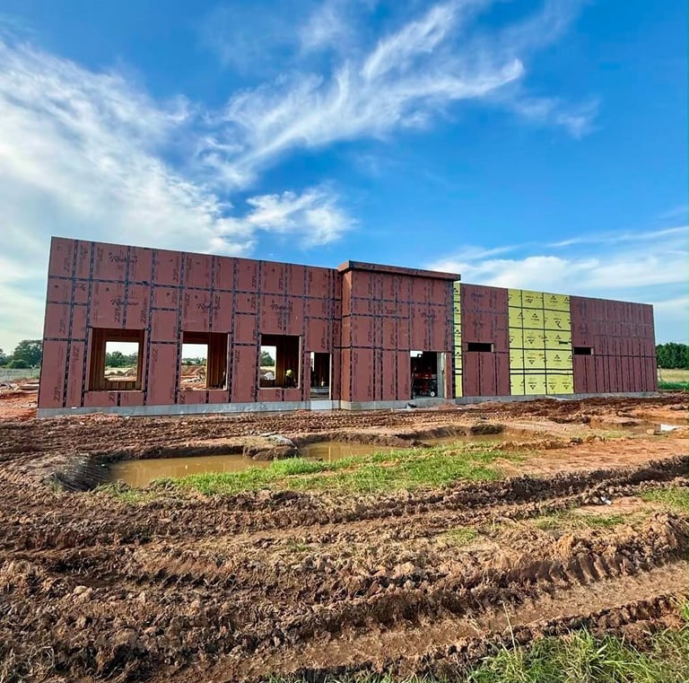 Modern barn under construction with metal siding and green accents on rural dirt foundation against clear blue sky