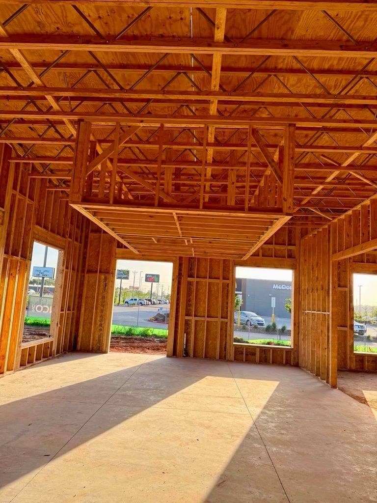Interior of an unfinished wooden house frame under construction showing exposed beams, trusses, and multiple windows with concrete floor